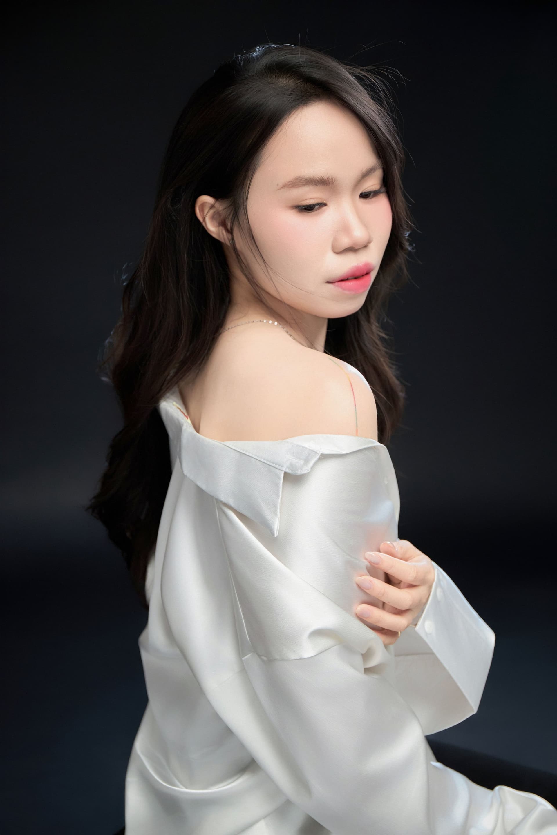 Cô gái áo trắng ngồi cúi nhẹ, lưng quay nửa mặt, ánh sáng dịu trên nền đen — khoảng lặng tuổi đôi mươi giữa Sài Gòn