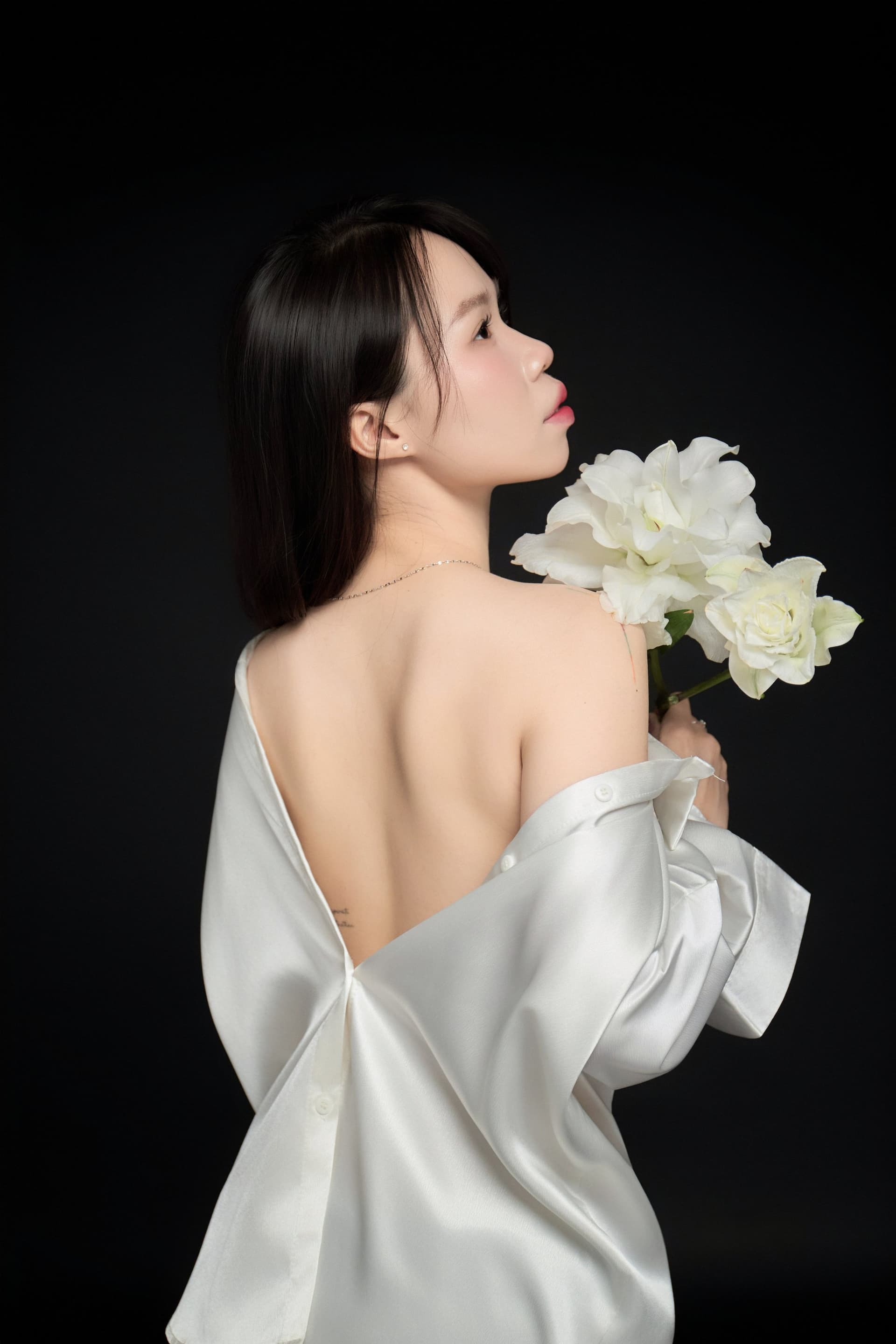 Cô gái áo trắng quay lưng, vai trần nhẹ trên nền tối — dáng đi tới của một mùa chưa chịu khép lại