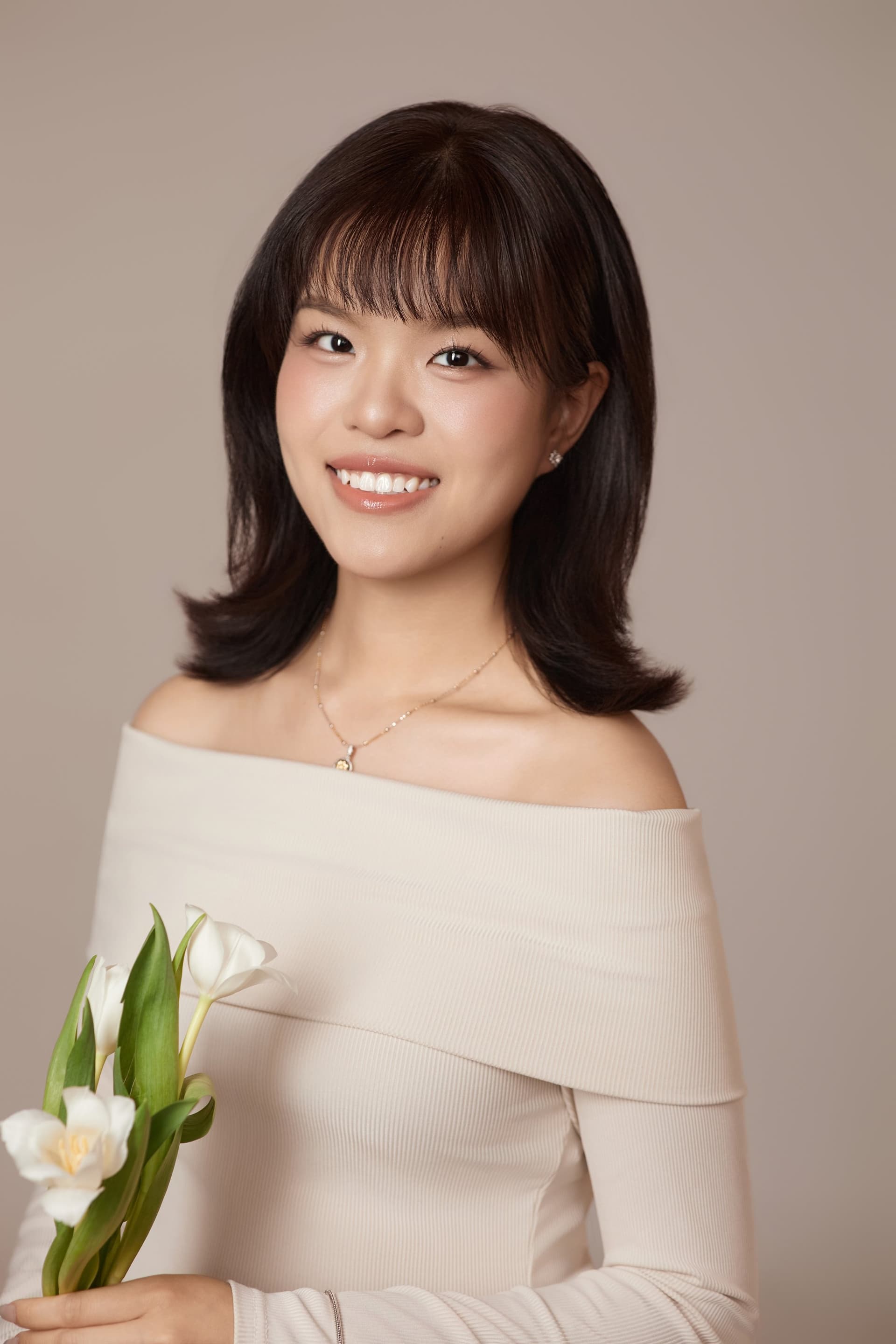 Người phụ nữ áo trắng trễ vai, đứng cầm hoa tulip trắng cuống dài, ánh nhìn nhẹ, nền nâu ấm — Ngọc trong khoảng riêng của mình
