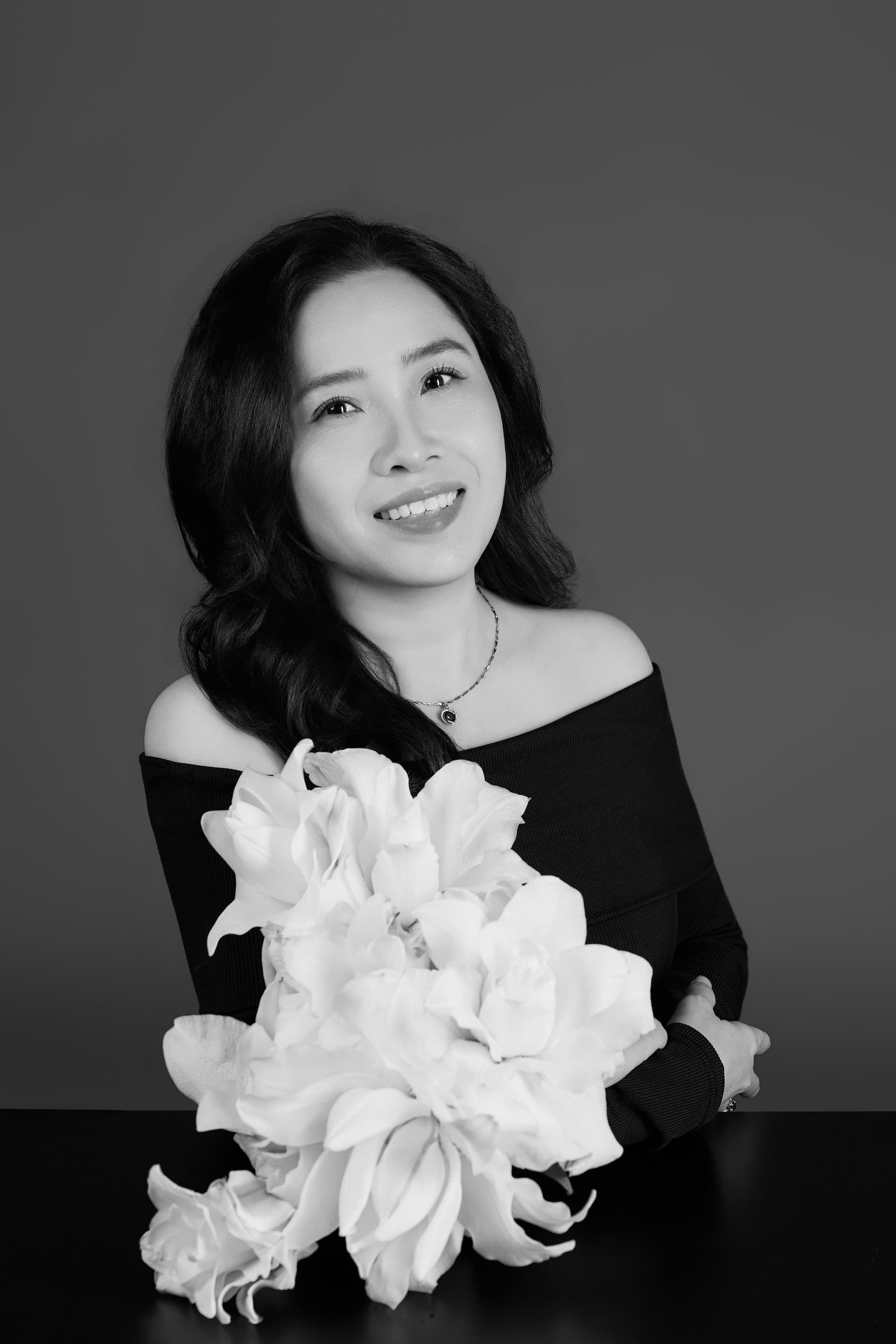 Chị Thin Thin mặc đầm đen trễ vai ôm bó hồng trắng dịu dàng khẽ mỉm cười tự nhiên