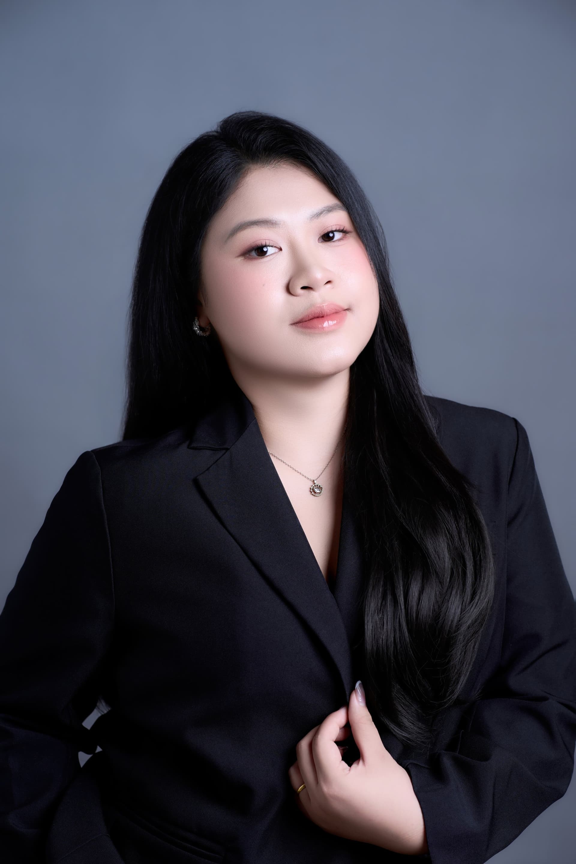 Người phụ nữ áo đen, tóc buông dài, gương mặt chuyên nghiệp trên nền tối — dáng vẻ của một giáo viên đang tự đứng riêng