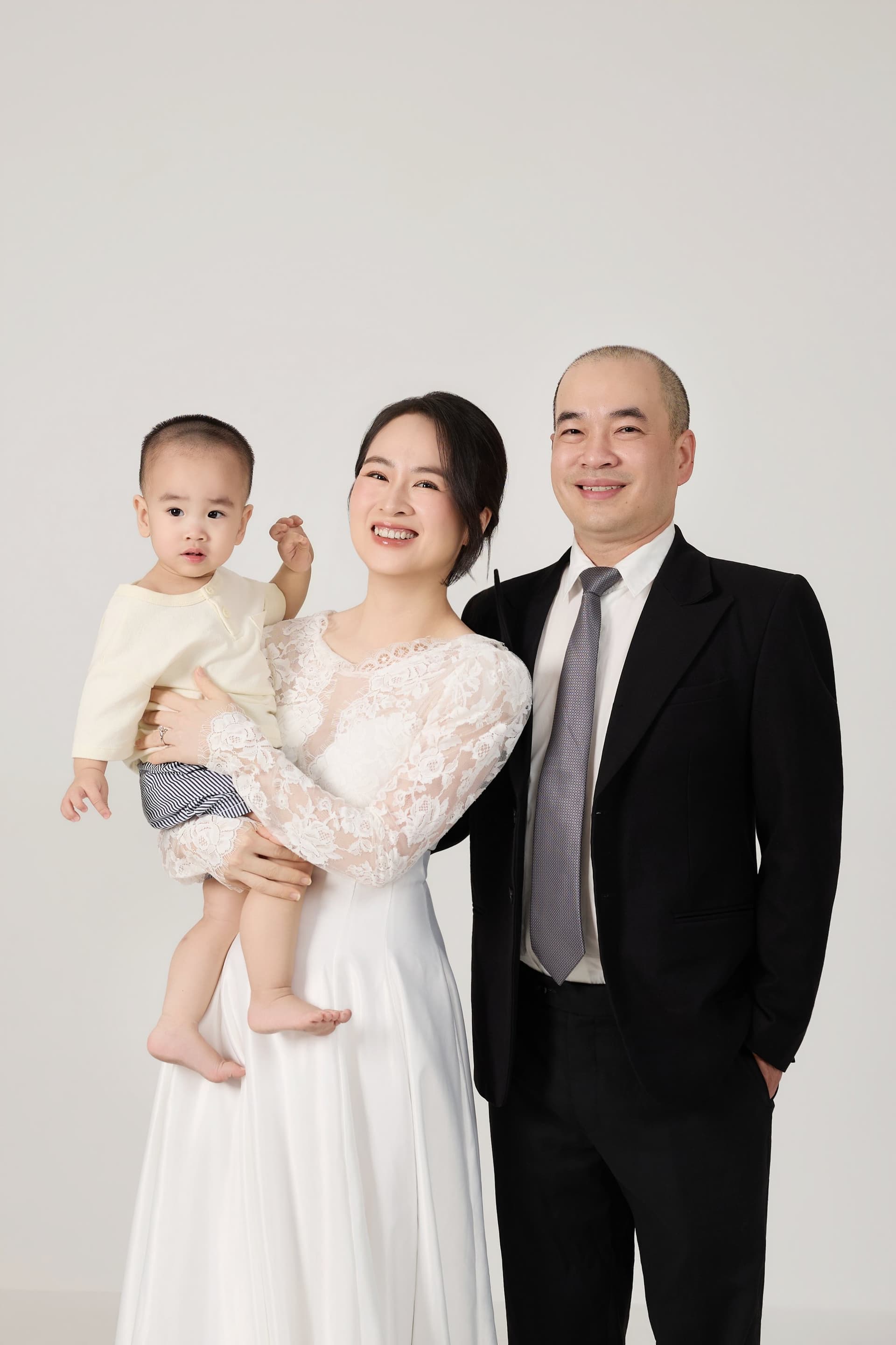 Hai vợ chồng mặc đồ cưới bế đứa con nhỏ ôm chặt cổ mẹ tràn ngập tình thương