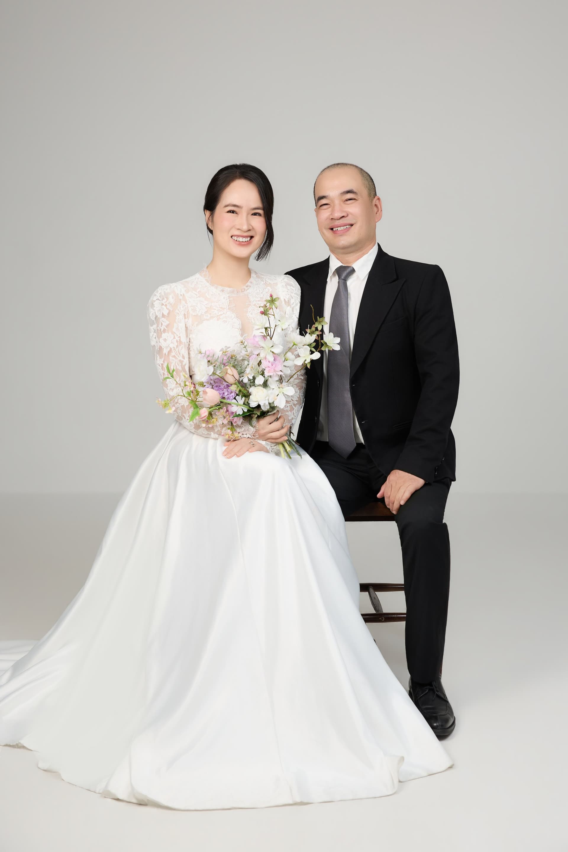 Chú rể mặc vest đen ngồi tựa vào cô dâu váy cưới trắng tinh khôi dưới ánh nắng nhẹ