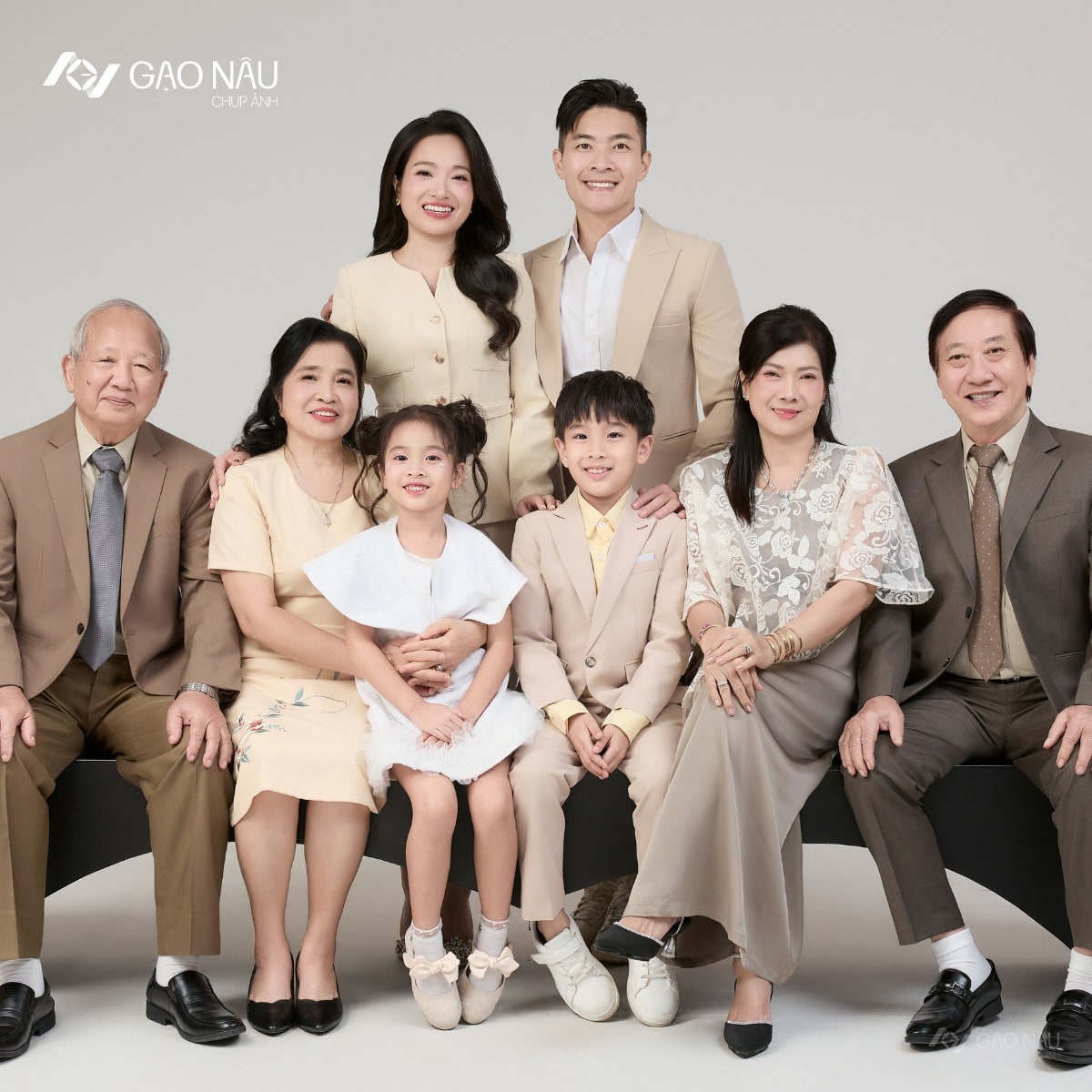 Đại gia đình tám người ba thế hệ đứng quây trong studio — ông bà nội, ông bà ngoại, vợ chồng trẻ đứng sau, hai cháu nhỏ đứng giữa, tone trang phục be nâu
