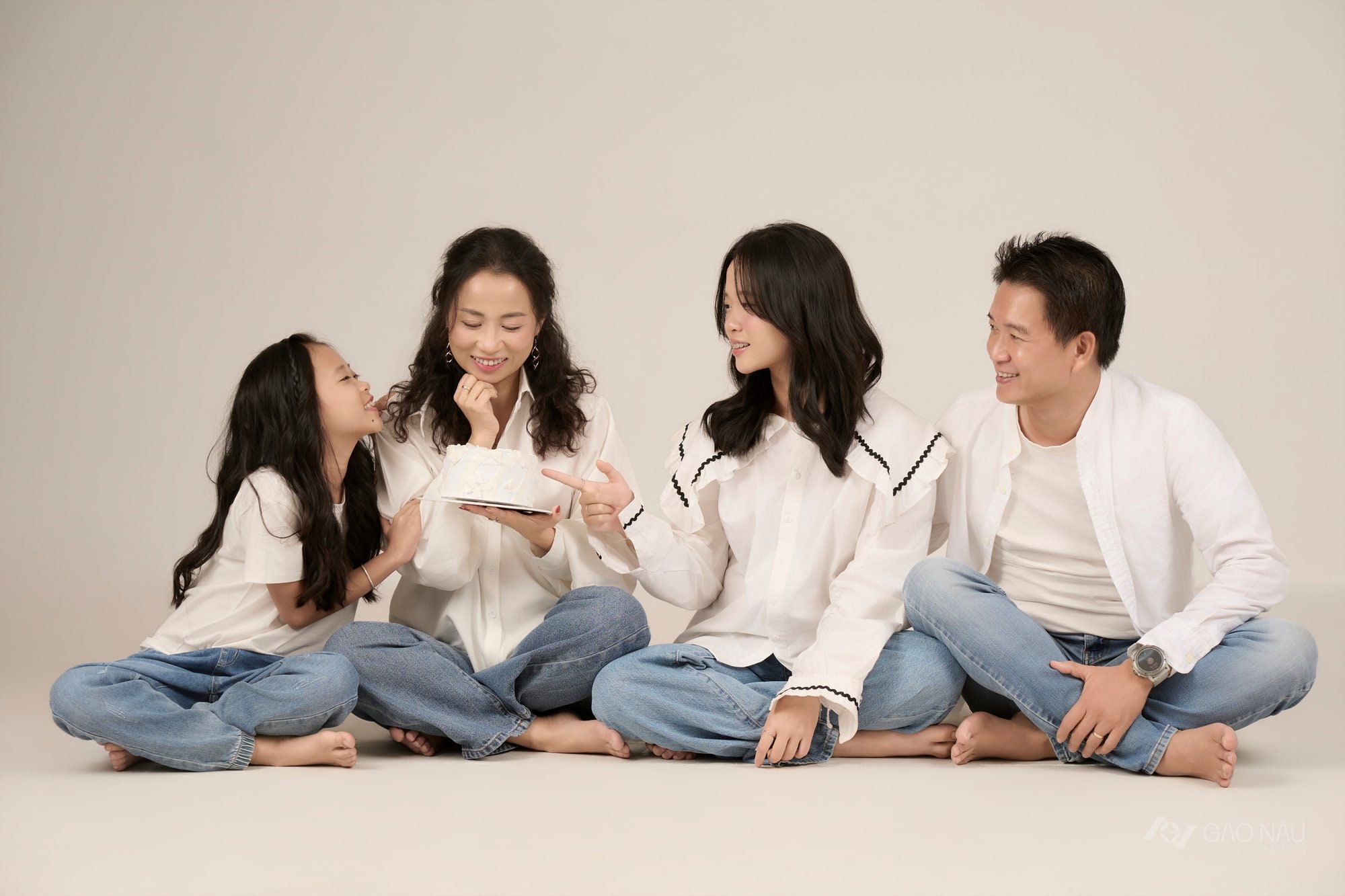 Gia đình bốn người ngồi quây quần trên sàn quanh chiếc bánh sinh nhật, chồng bên trái, vợ bên phải, hai con gái ngồi giữa, không khí ấm cúng