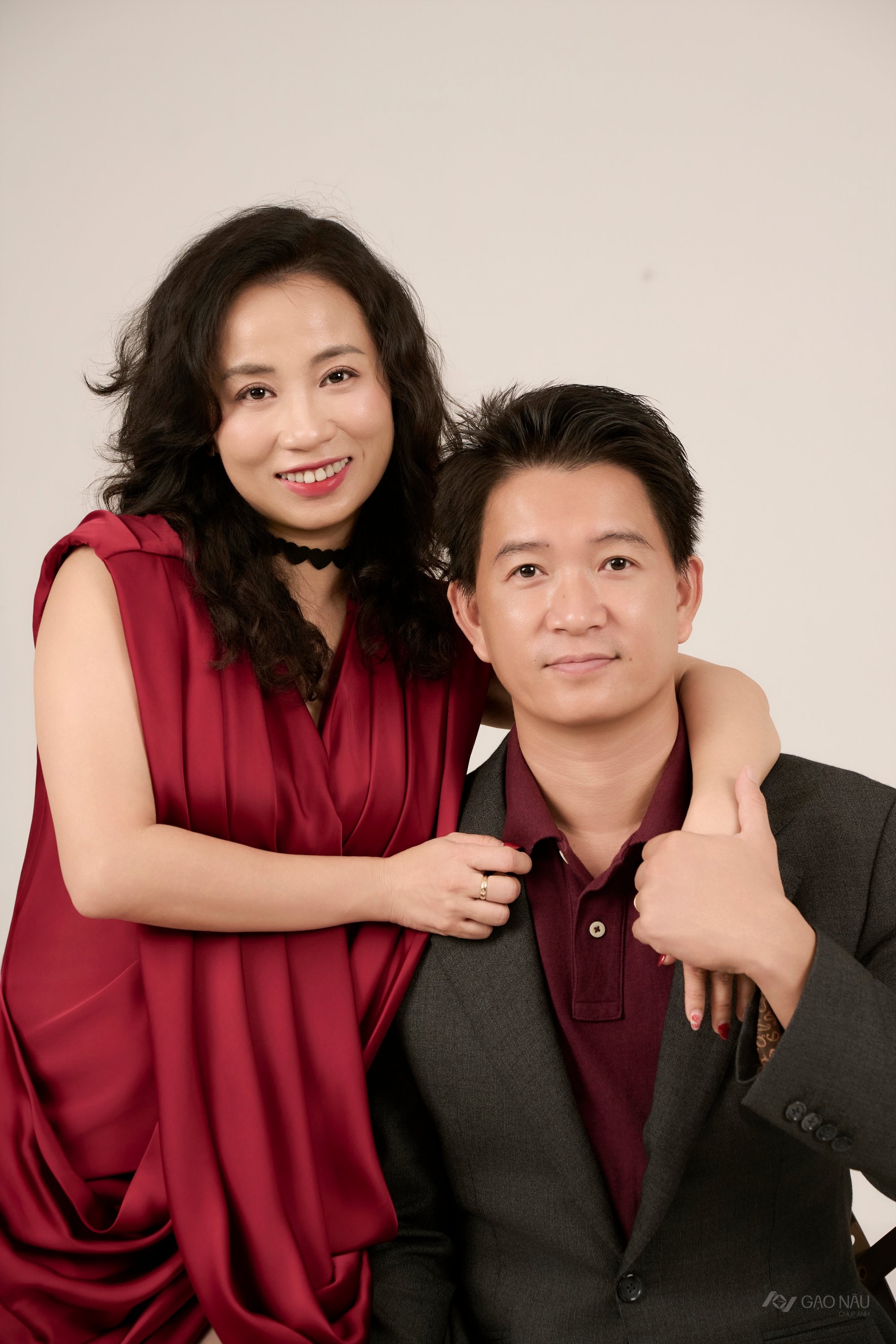 Vợ chồng đứng cạnh nhau — vợ áo lụa đỏ, chồng vest xám với áo polo burgundy bên trong, cả hai nhìn về phía trước, nền ấm — kỷ niệm hai mươi sáu năm yêu nhau từ thời cấp ba