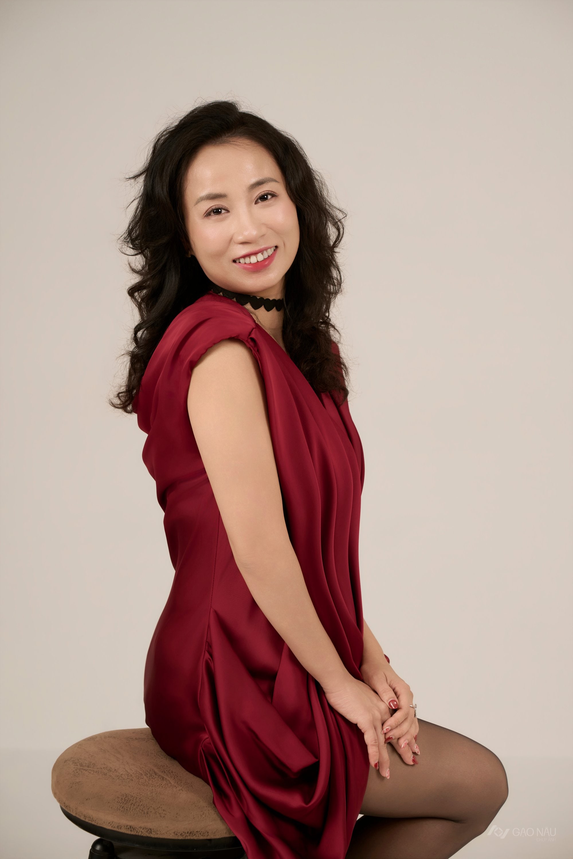 Người phụ nữ 38 tuổi ngồi trên ghế, áo dài lụa đỏ, tóc búi nhẹ, biểu cảm trầm tĩnh, nền trầm — hai mươi sáu năm vừa giữ một chỗ ngồi ở công ty vừa giữ một mái nhà
