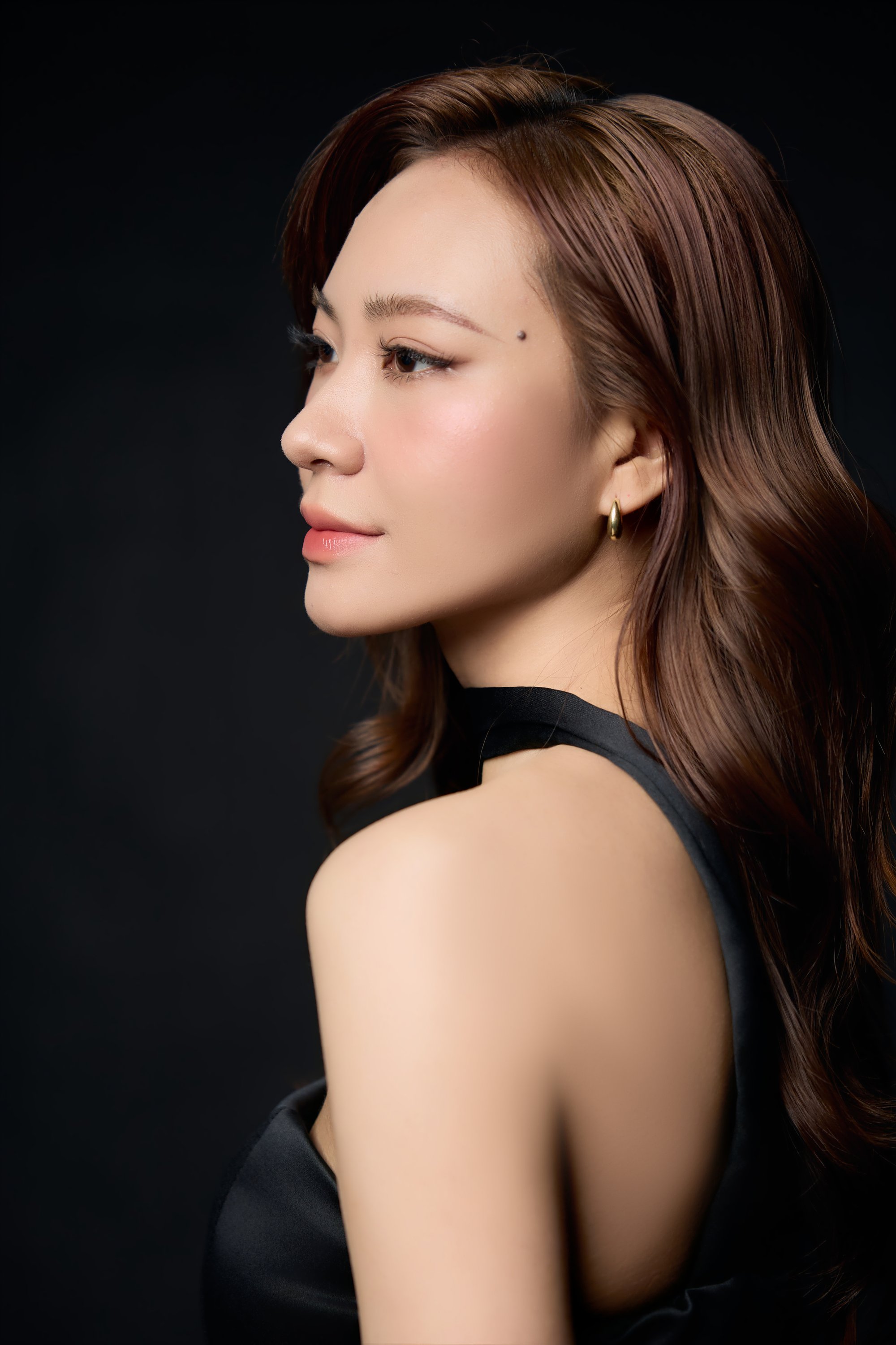 Khánh Trang quay nghiêng, vai trần, choker đen mảnh, gương mặt nhẹ nhõm trên nền tối — một người ít nói quen đứng ở phần quan sát
