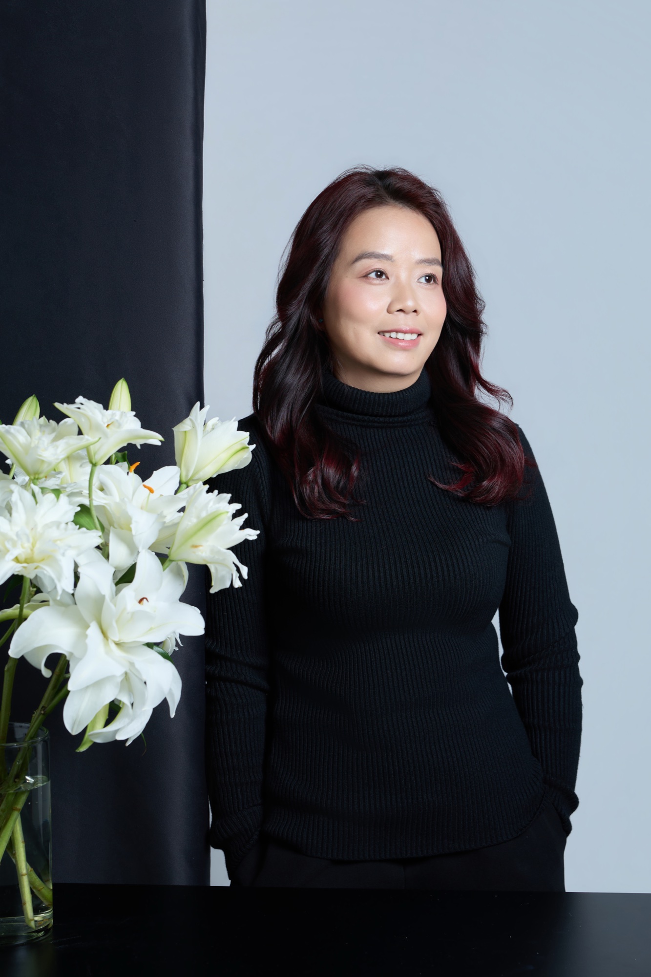 Người phụ nữ ngoài 40 mỉm cười nhẹ, đứng cạnh bình hoa loa kèn trắng, áo cao cổ đen, biểu cảm bình yên