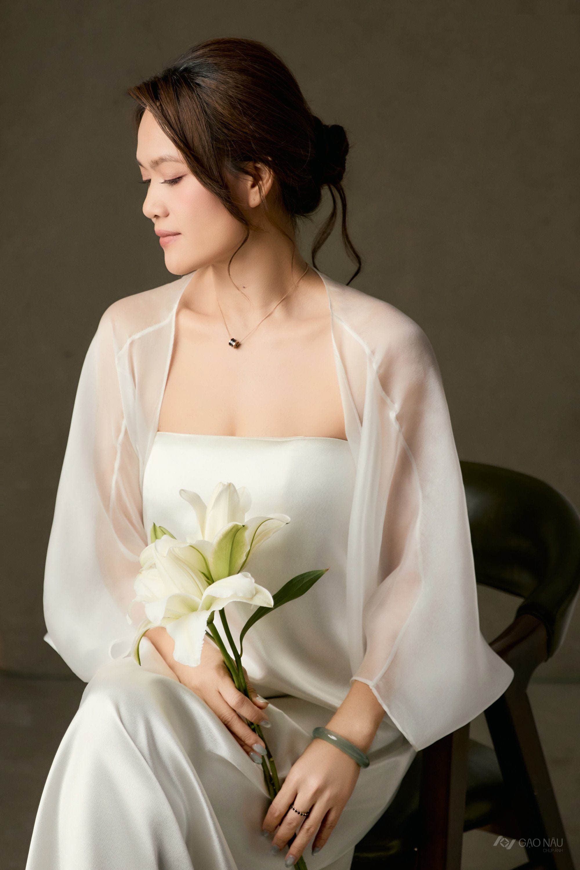 Người phụ nữ búi tóc lỏng, áo voan trắng, nhắm mắt nghỉ, bó loa kèn trắng đặt trên lòng, vòng ngọc bích trên cổ tay, nền nâu ấm — khoảnh khắc chạm của câu chuyện