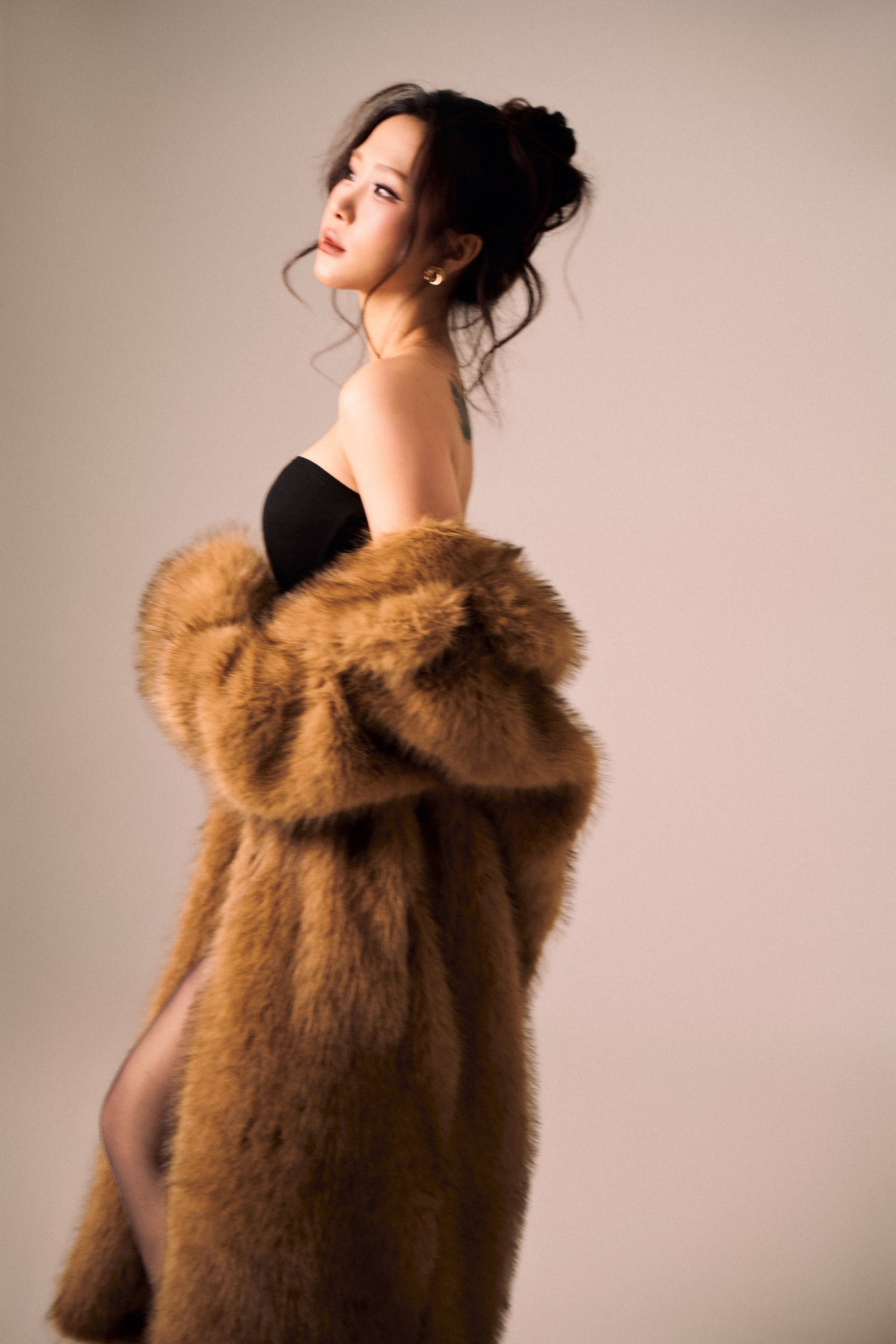 Thiếu nữ búi tóc lỏng, đứng nghiêng, khoác áo lông màu nâu vàng phủ ngoài đầm đen, ánh mắt chếch lên nhẹ — bản dạng "áo lông sắc"
