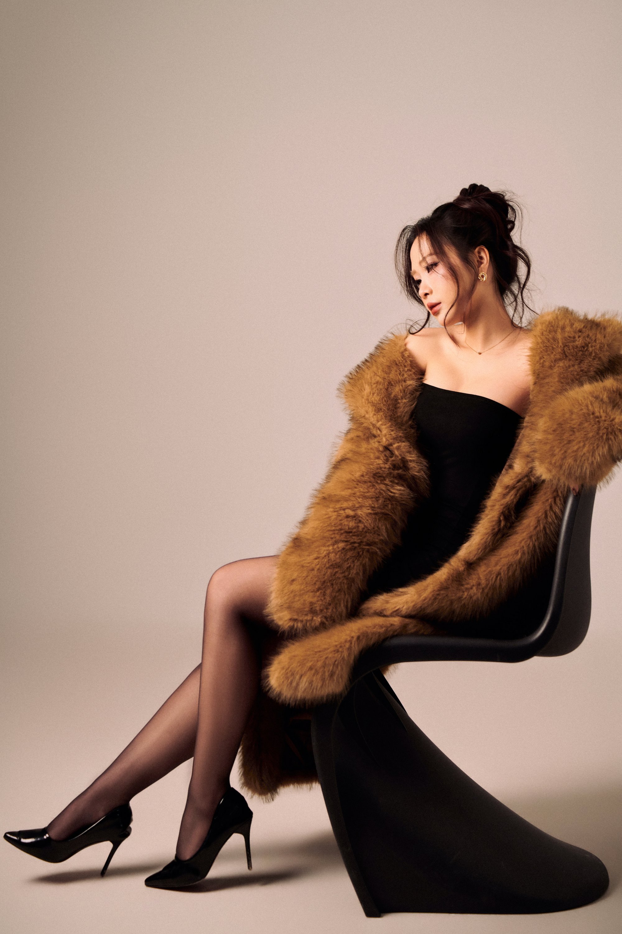 Thiếu nữ búi tóc cao, áo lông nâu vàng phủ vai, đầm đen, vắt chân ngồi, giày cao gót đen, ánh mắt nhìn xuống — sự đối lập của bản dạng "online"