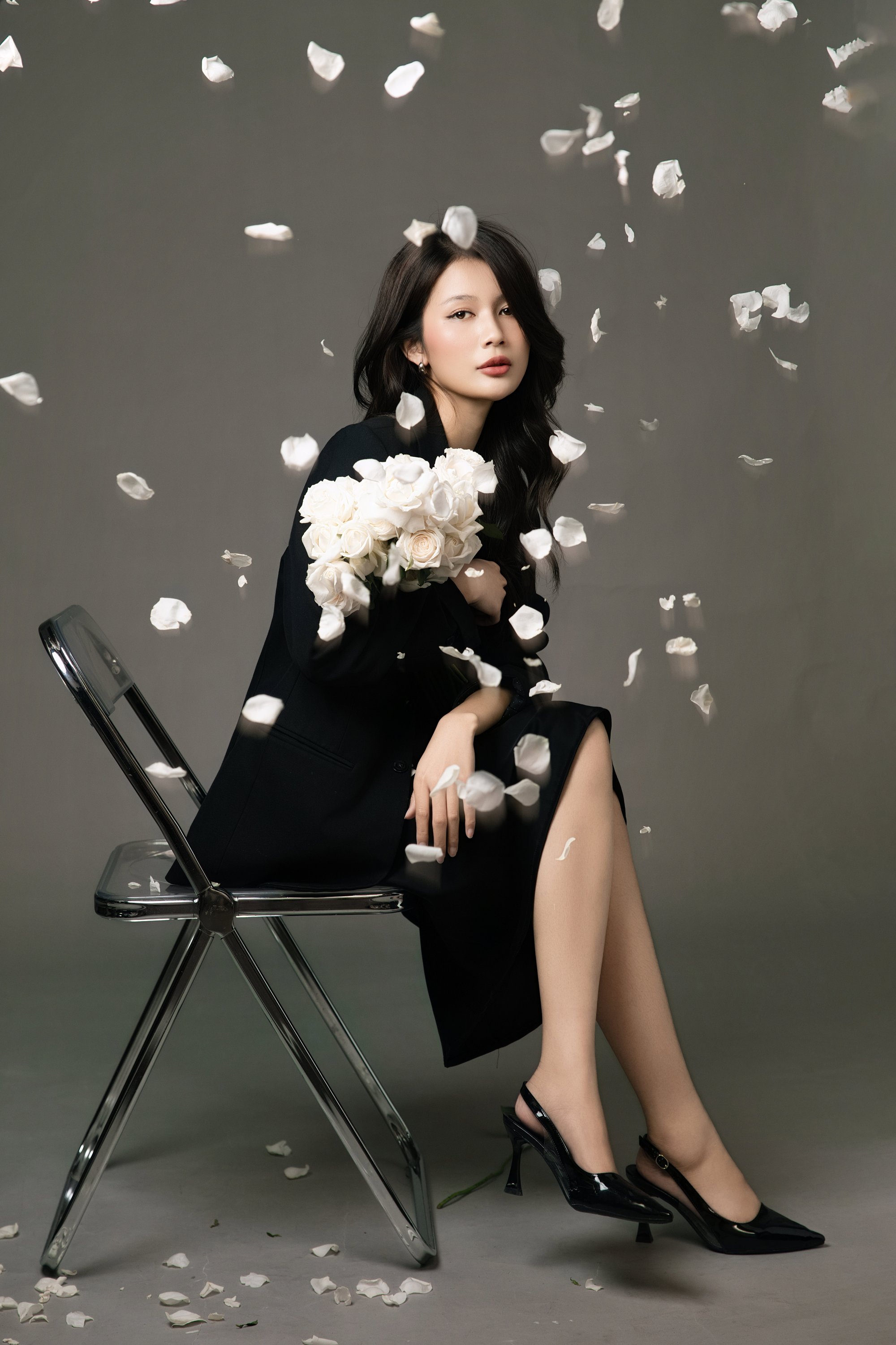 Người phụ nữ trong blazer đen ngồi ghế gấp giữa nền sage, cánh hoa rơi quanh sàn — toàn cảnh tĩnh, một mình
