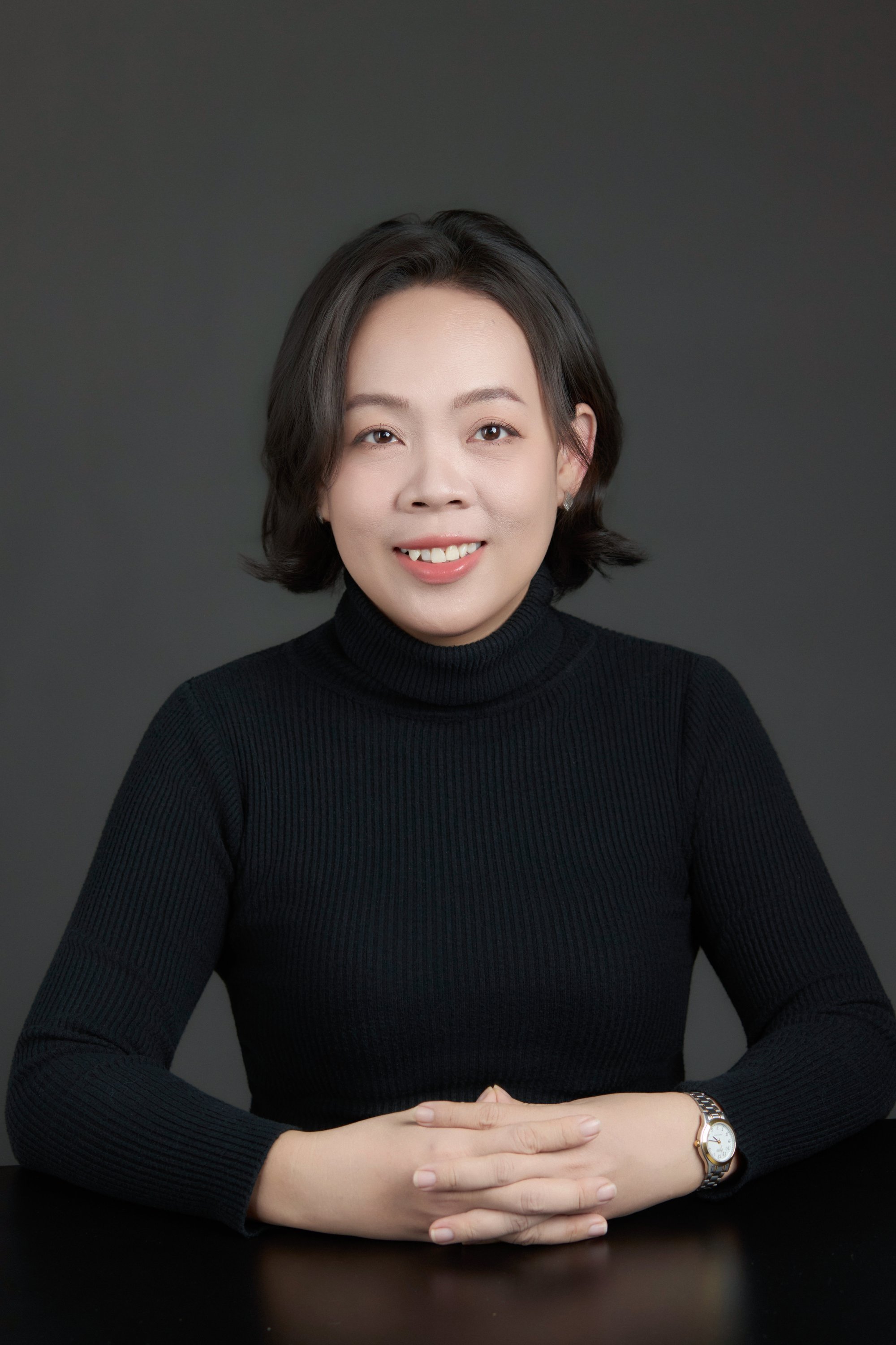 Bán thân — người phụ nữ tóc ngắn, áo cổ lọ đen, hai tay đan vào nhau đặt trên bàn, đeo đồng hồ kim loại trên cổ tay trái — tư thế của một người ngồi đối diện ai đó để lắng nghe