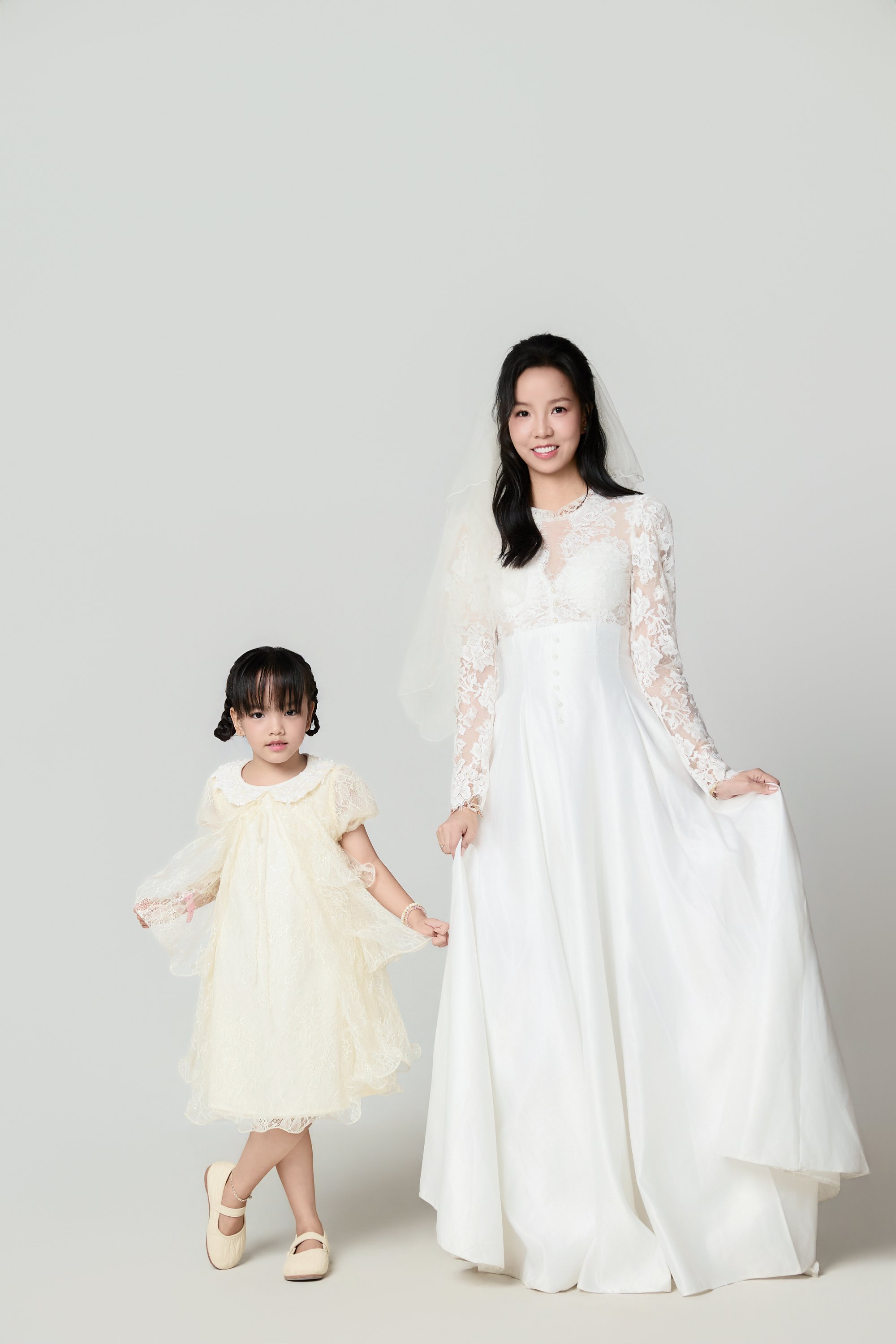 Người mẹ trong váy cô dâu trắng và con gái nhỏ váy trắng đứng đối diện nhau giữa nền dịu — hai vợ chồng cùng nhìn về một hướng, hướng đó là con